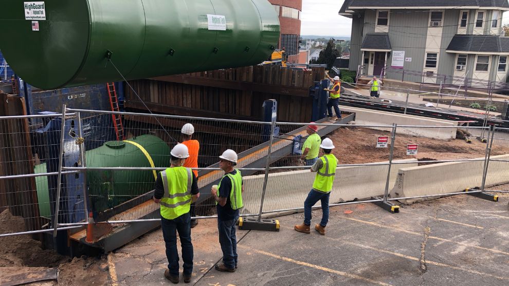 Construction workers in hard hats and safety vests watch as the new underground storage tank is installed